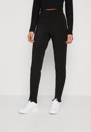 Czarne spodnie wysoka talia o dopasowanym kroju, subtelne rozcięcia po bokach przy kostce, w połączeniu z białymi trampkami. Gładka faktura i eleganckie linie.