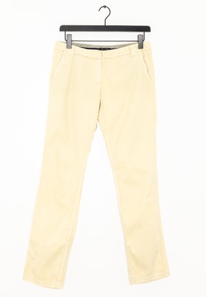 Beżowe spodnie o prostych nogawkach z pętlami na pasek i bocznymi kieszeniami, zawieszone na czarnym wieszaku na ubrania na białym tle.