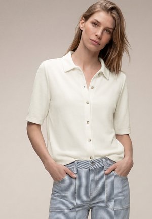 Kobieta o jasnobrązowych włosach ubrana w białą koszulę na krótki rękaw zapinaną na guziki oraz niebieskie spodnie w paski, stoi z rękami w kieszeniach na jednolitym tle.