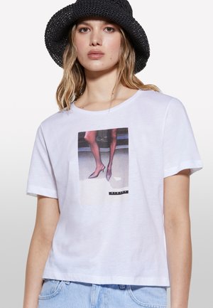 T-shirt bianco in cotone con design accorciato, caratterizzato da una stampa grafica di gambe in tacchi alti. Maniche corte e vestibilità rilassata.