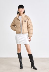 Doudoune beige, texture matelassée, fermeture éclair, col haut, avec un logo 'ck' marron. Associée à une robe blanche ajustée et des bottes noires montantes.