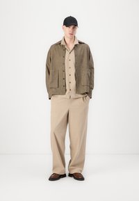 Brown checkered jacket with two front pockets, beige button-up shirt underneath, beige wide-leg pants, and a black cap, standing on a white background.