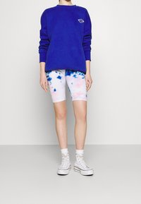 Modrá mikina s malým logem, bílo-růžové tie-dye cyklistické šortky a bílé tenisky s vysokým svrškem a kotníkovými ponožkami. Jednoduchý, ležérní outfit.