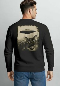 Black sweatshirt featuring a cat's face with large eyes, surprised expression, and a UFO above, all in a distressed beige print.
