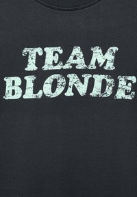 Tessuto nero con testo azzurro chiaro in stile vintage che recita "TEAM BLONDE" in grassetto, centrato su di esso.