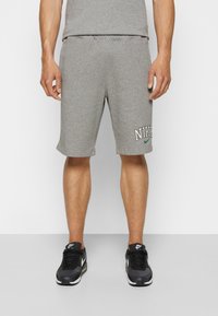 Muž v šedých Nike šortkách s logem na levé noze a černých teniskách Nike Air Max, stojící na jednolitém pozadí.