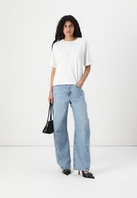 Hvit kortermet t-skjorte kombinert med lys blå vide jeans og svarte spisse hæler, holder en liten svart håndveske.