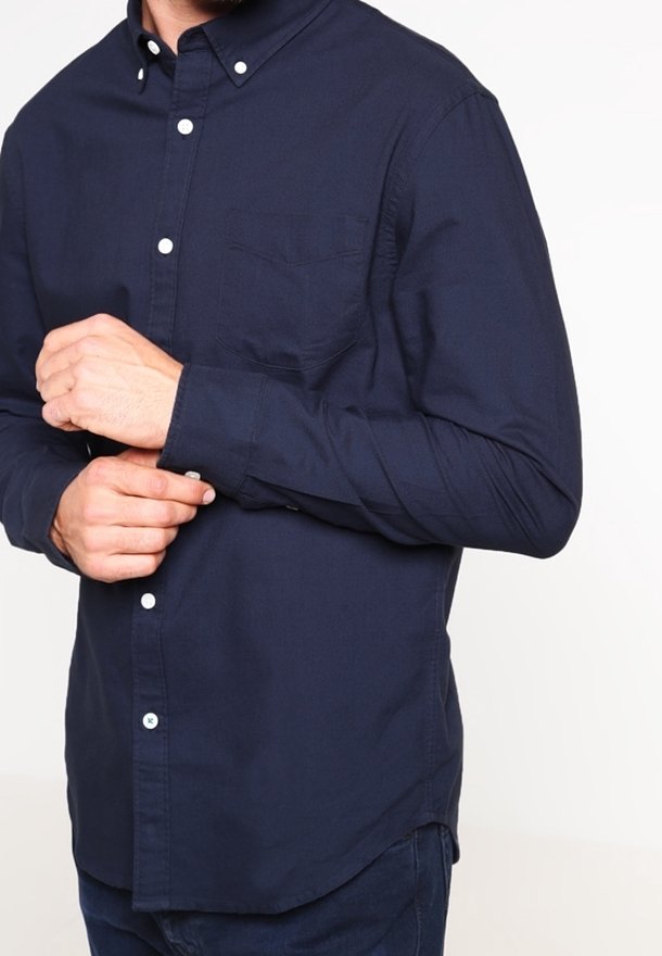 Mörkblå långärmad skjorta med knappar, bröstficka, vita knappar och spetsig krage. Tyget verkar slätt och åtsittande.