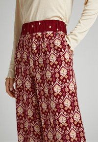 Breda byxor i röd och krämfärgad mönsterdesign med geometriska former. Elastisk midja, sidofickor och lättviktsmaterial.