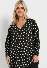 Svart blus med stora krämfärgade prickar, V-ringning, långa puffärmar och texturerade manschetter. Tyget har en slät finish.