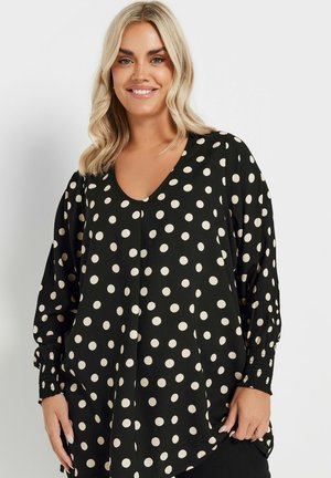 Blusa nera con grandi pois crema, scollatura a V, maniche lunghe a sbuffo e polsini texturizzati. Il tessuto ha una finitura liscia.