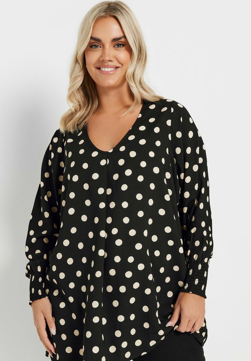 Svart blus med stora krämfärgade prickar, V-ringning, långa puffärmar och texturerade manschetter. Tyget har en slät finish.