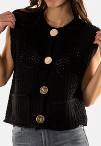 Chaleco negro de punto con un patrón texturizado, grandes botones redondos dorados y dos bolsillos delanteros. Diseño sin mangas, ajuste casual.