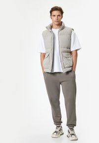 Chaleco acolchado gris claro con un alto cuello y dos bolsillos frontales, usado sobre una camiseta blanca y combinado con pantalones de chándal grises y zapatillas con estampado.