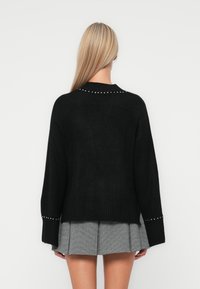 Pull en tricot noir avec ourlet côtelé et manches larges, orné de clous argentés le long du haut du dos et des poignets. Assorti à une jupe plissée grise.