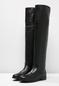 Une paire de bottes en cuir noir hautes avec des talons bas et des orteils ronds, se tenant droites sur une surface blanche, sur un fond clair et uni.