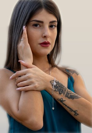 Silberarmband mit Herzanhänger und filigranem kreisförmigen Design, getragen an einem Handgelenk mit einem detaillierten Tattoo, vor einem sanften Hintergrund.