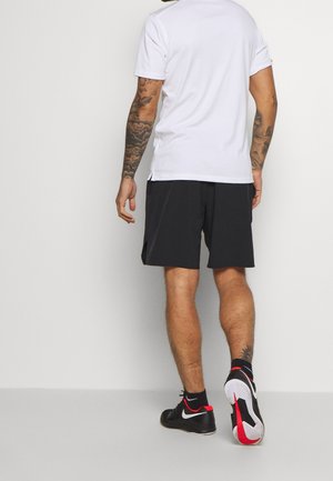 Mand med tatoveringer iført en hvid T-shirt, sorte shorts og sorte Nike-sneakers, der går væk på en ensfarvet lys baggrund.