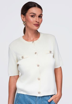 Femme aux cheveux bruns en queue de cheval basse, portant des boucles d’oreilles en or et un haut blanc à manches courtes boutonné avec poches à rabat, posant avec la main dans la poche.