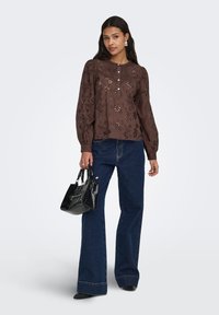 Braune, bestickte Bluse mit langen Ärmeln, kombiniert mit hochtaillierten blauen Schlagjeans. Hält eine kleine schwarze Handtasche und trägt spitze schwarze Schuhe.