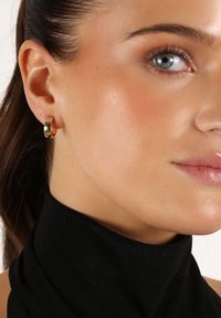 Guldörhängen med polerad yta, cirkulär form och slät textur. Bärs av en person med svart topp och minimal makeup.