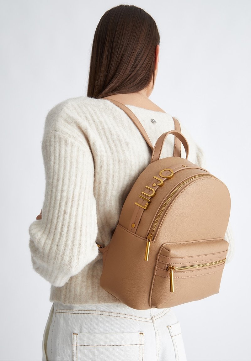 Femme aux longs cheveux bruns portant un pull blanc et un pantalon clair, avec un sac à dos en cuir beige doté de fermetures éclair dorées et d’un détail du nom de la marque.