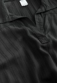 Chemise en tissu noir texturé avec un col en V et une étiquette intérieure indiquant "NEXT" et "downtime".