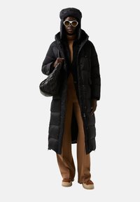 Long manteau noir matelassé avec capuche, associé à un haut marron et un pantalon large. Les accessoires comprennent un sac noir et des lunettes de soleil rondes blanches.