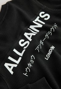 Čierna textilná odev s vyvýšenou textúrou, s tučnými bielymi nápismi "ALLSAINTS" a "LONDON" spolu s japonskými znakmi.