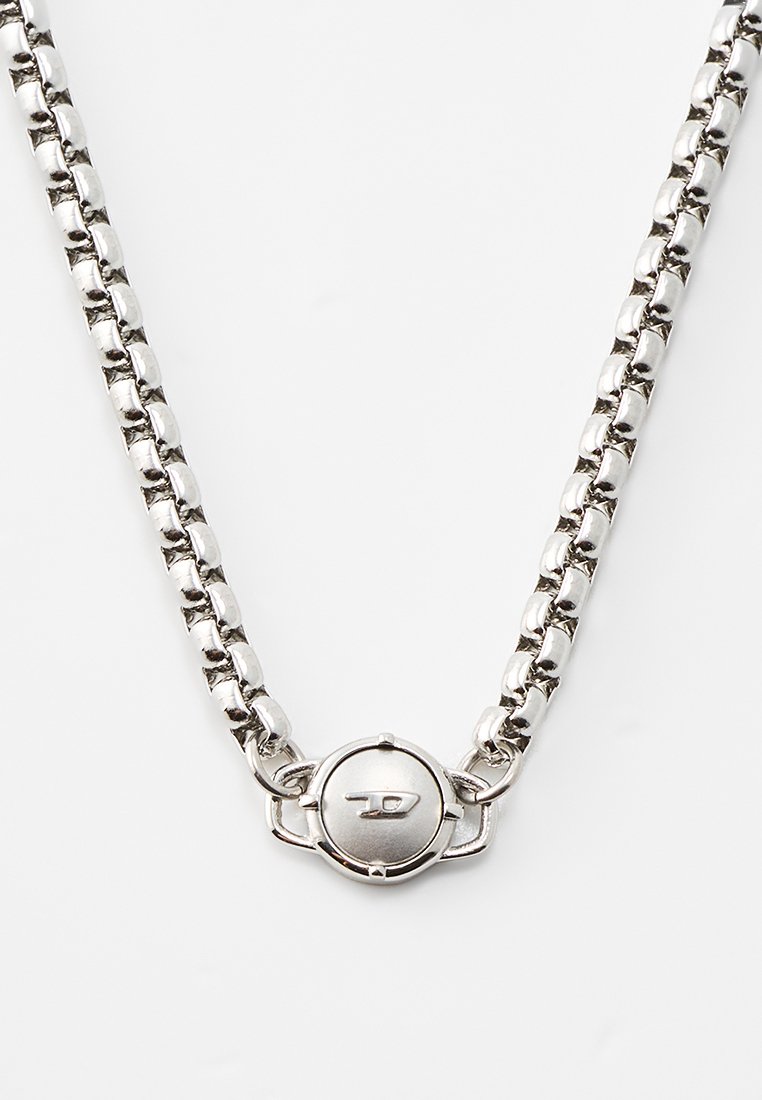 Collier en chaîne argentée avec des maillons ronds, présentant un fermoir circulaire avec un logo. Texture lisse avec une finition polie.