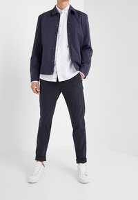 Mies, joka on pukeutunut tummansiniseksi nappivaatteeseen, valkoiseen paitaan, tummiin käärittyihin housuihin ja valkoisiin tenniskenkiin, seisoo tasaisella valkoisella taustalla.