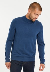 Kobaltblauer gestrickter Pullover mit einem hohen, gerippten Kragen, langen Ärmeln und einem geraden Saum, der ein kleines Logotag am unteren Rand aufweist.