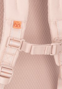 Beige hátizsák pántok állítható műanyag csatokkal, textúrált szövet háttérrel és narancssárga logó címkével, amely grafikus designnal rendelkezik.