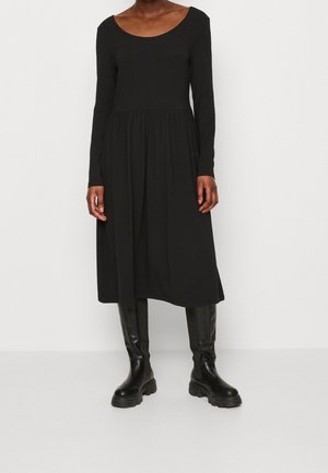 Nainen pukeutuneena pitkähihainen musta midimekko ja polven korkeuteen yltävät mustat paksupohjaiset saappaat, seisomassa yksinkertaista valkoista taustaa vasten.