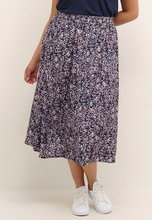 Kvinde iført en marineblå midi-nederdel med blomsterprint og lyserøde og hvide prikker, kombineret med hvide sneakers og en marineblå top, stående mod en enkel baggrund.