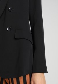 Czarna dopasowana marynarka z dwoma guzikami, klapami wyłogowymi i jedną kieszenią na piersi. Materiał wydaje się gładki z dopasowanym fasonem.