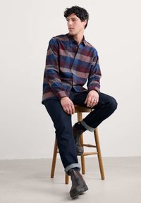 Camicia a quadri in tonalità blu scuro, rosso e grigio, con bottoni e una tasca sul petto. Indossata con jeans scuri e stivaletti marroni. Seduto su uno sgabello di legno.