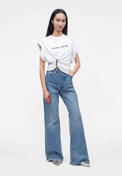 Femme debout, portant un t-shirt blanc Marc Jacobs cintré à la taille, un jean bleu à jambes larges et des chaussures pointues argentées sur fond uni.
