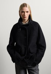 Cappotto nero dalla texture con colletto alto, dotato di grandi bottoni e due tasche frontali, abbinato a jeans scuri. Design semplice e strutturato.