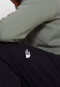 Osoba oblečená v šedé košili s dlouhým rukávem a černých kalhotách s vyšívaným logem The North Face na stehně.