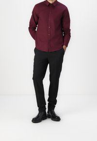 Mężczyzna w burgundowej koszuli z guzikami, czarnych spodniach i czarnych skórzanych butach stoi na białym tle.