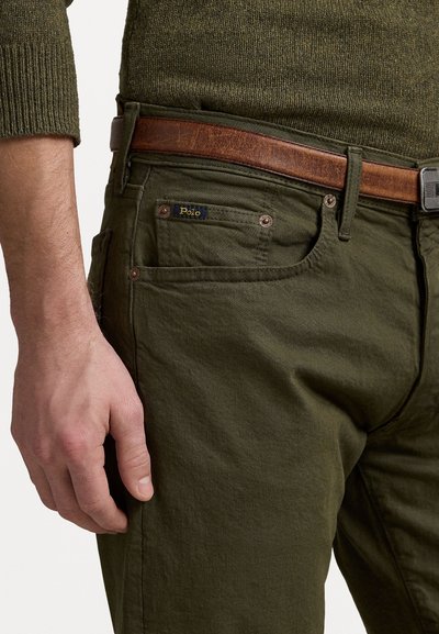 Ελαιοπράσινα βαμβακερά chinos με καφέ δερμάτινη ζώνη. Περιλαμβάνει μπροστινές τσέπες και μια μικρή μπλε ετικέτα "Polo" στην πίσω τσέπη.