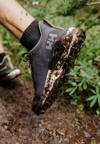 Běžecká obuv na trail v černé síťovině se zelenými akcenty, pokrytá blátem. Výrazná texturovaná podrážka pro přilnavost na nerovném terénu.