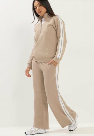 Femme debout portant un survêtement beige avec des bandes blanches sur les côtés, des baskets blanches et un sweat à capuche zippé, mains dans les poches.