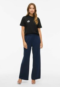 Zwarte katoenen t-shirt met een lippenprint, gecombineerd met marineblauwe wijde broek. Het model draagt doorzichtige hakken, wat een casual uitstraling geeft.