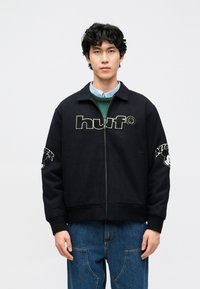 Casaco preto com fecho de correr, com logótipo "huf" bordado na frente e desenho do Mickey Mouse na manga esquerda. Tecido macio, punhos canelados.