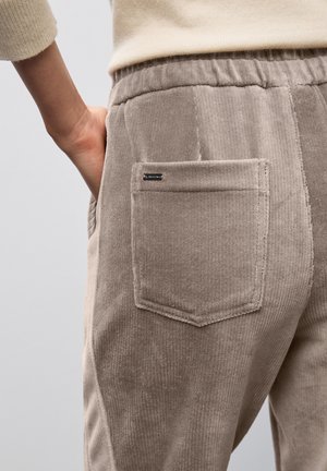 Persona che indossa pantaloni in velluto a coste beige con vita elastica, mano posizionata nella tasca laterale e tasca posteriore visibile.