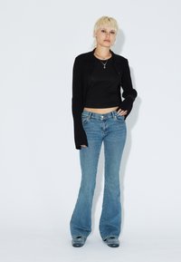 Svart ribbad cardigan över en svart crop top, med blå flare jeans. Strukturerad tyg, figurnära silhuett och medelhög midja.