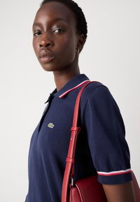 Tamsiai mėlynas polo marškinėliai su raudona ir balta juostele prie apykaklės, kuriuose yra mažas žalias Lacoste logotipas. Raudona rankinė mėgstama nešioti ant peties.