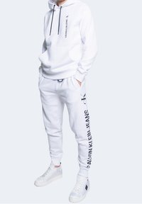 Sudadera con capucha y pantalones de chándal blancos, con texto negro "CALVIN KLEIN JEANS". El material parece suave; el calzado es blanco con suela texturizada.